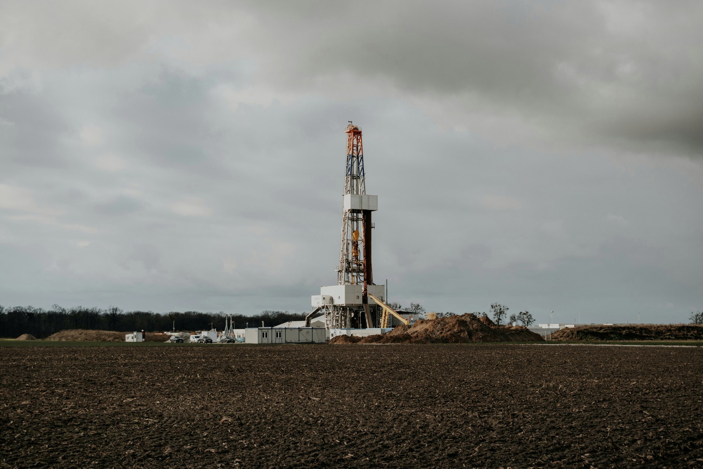A drilling rig standing at the edge of a plowed field under a gray sky
